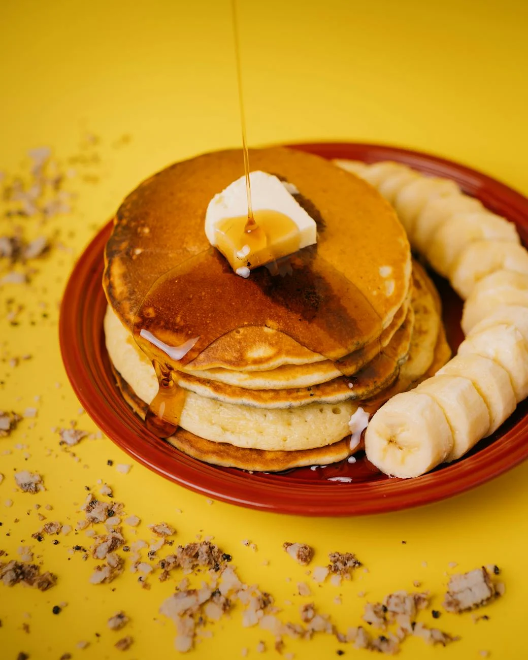 Stack of pancakes with syrup and banana slices on a vibrant yellow background.